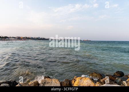 Vue panoramique sur la côte de la mer Noire avec des vagues frappant les rochers, la plage et l'horizon de la ville côtière sous la lumière douce du soir. Banque D'Images