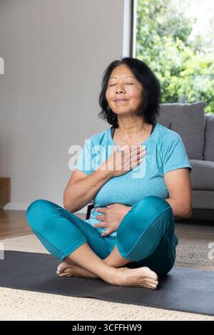 Femme asiatique senior portant des vêtements de sport pratiquant la respiration sur tapis de yoga dans le salon à côté du canapé Banque D'Images