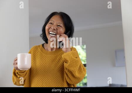 Femme asiatique senior souriant tout en tenant smartphone et tasse blanche à la maison portant un pull moutarde Banque D'Images