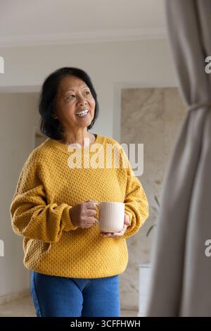 Femme asiatique senior tenant une grande tasse en céramique souriante regardant droit dans le salon portant un pull Banque D'Images