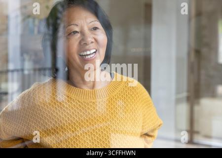 Femme asiatique senior debout derrière un panneau de verre à la maison portant un pull moutarde réfléchissant la lumière Banque D'Images
