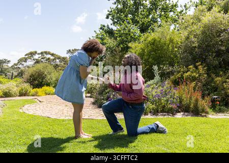 Couple afro-américain agenouillé dans un jardin ensoleillé sur un chemin de briques courbé, l'homme proposant Banque D'Images