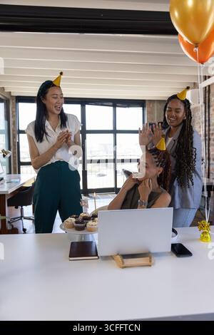 Diverses collègues féminines portant des chapeaux de cône célébrant l'anniversaire au bureau avec cupcakes smartphone Banque D'Images