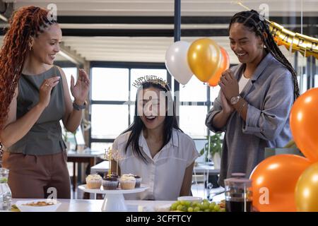 Diverses collègues féminines célèbrent leur anniversaire dans la zone de pause du bureau avec des cupcakes et une bannière dorée Banque D'Images