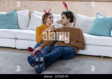 Couple assis à la maison portant des bandeaux en bois et des chaussettes tricotées tenant des tasses en céramique près d'un mur de bois Banque D'Images