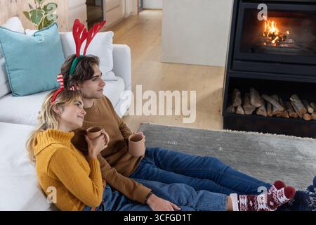 Couple portant des bandeaux en bois rouge appuyé contre le canapé, tenant des tasses à la maison devant la cheminée Banque D'Images