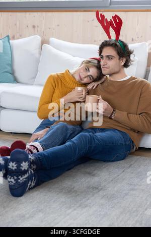 Couple assis sur un tapis gris dans le salon tenant des tasses beiges et portant des bandeaux en bois rouge Banque D'Images