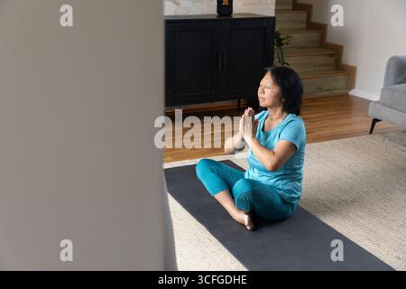 Femme senior asiatique méditant sur un tapis de yoga à la maison portant un haut turquoise, leggings sarcelle, espace copie Banque D'Images