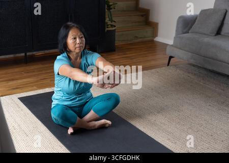 Femme asiatique d'âge moyen étirant les bras vers l'avant assis sur un tapis de yoga à la maison dans des vêtements de sport turquoise Banque D'Images