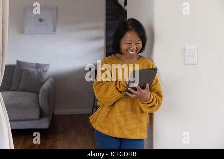 Femme asiatique sénior tapant la tablette portant le pull moutarde dans le salon par canapé, art, espace de copie Banque D'Images