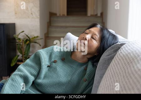 Femme asiatique senior portant un pull sarcelle dormant sur un canapé gris avec un oreiller gris clair, espace copie Banque D'Images