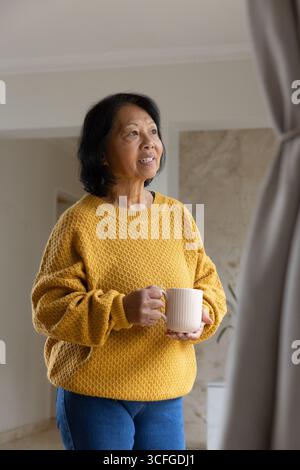 Femme asiatique senior portant chandail moutarde tenant la tasse et regardant par la fenêtre par la plante en pot Banque D'Images