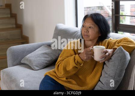 Femme asiatique senior assise sur le canapé avec tasse regardant à travers la fenêtre dans le salon, espace de copie Banque D'Images