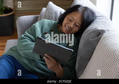 Femme asiatique senior s'inclinant sur le canapé à côté du panier de jardinière tissé tenant l'ordinateur de tablette Banque D'Images