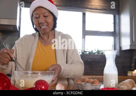 Femme asiatique senior portant chapeau de Père Noël mélangeant la pâte avec fouet, bol, œufs, lait au comptoir de cuisine Banque D'Images