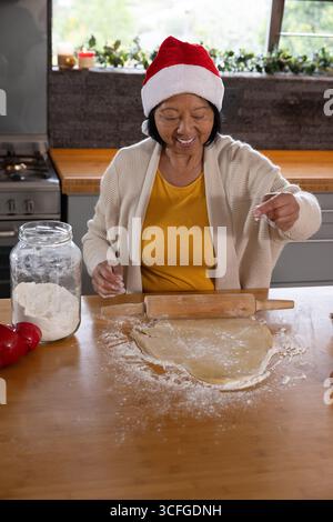 Femme asiatique senior portant chapeau de Père Noël pâte à rouler avec rouleau à pâtisserie et saupoudrer de farine dans la cuisine Banque D'Images