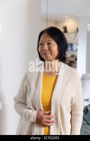 Femme asiatique senior debout à l'intérieur du salon à côté de la colonne, portant un cardigan beige, haut moutarde Banque D'Images