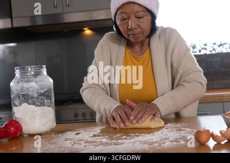 Femme asiatique senior portant chapeau de chef pétrissant la pâte au comptoir de cuisine avec pot de farine, coquilles d'œufs Banque D'Images