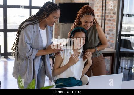 Diverses collègues féminines se penchant et souriant autour de l'ordinateur portable sur le bureau avec la plante en pot dans le bureau Banque D'Images