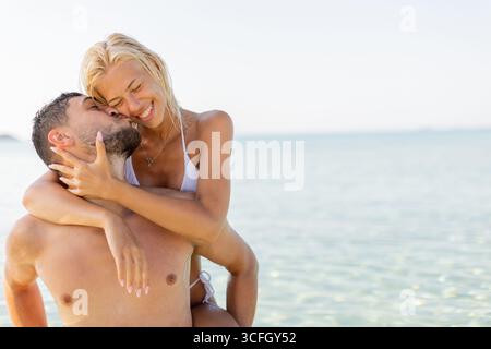 Joyeux couple partage une étreinte intime à la plage, souriante et la vie aimante par l'eau sereine sur une journée ensoleillée brillante. Banque D'Images
