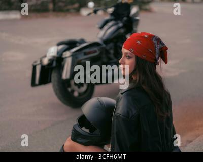 Jeune femme motard élégante dans une veste en cuir et bandana rouge est assis sur l'escalier avec son casque de moto près de son vélo. Banque D'Images