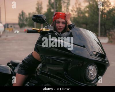 Jeune femme motard élégante en veste en cuir et bandana rouge est assis en toute confiance sur une moto, regardant la caméra. Urbain cool, dynamique et rebelle Banque D'Images
