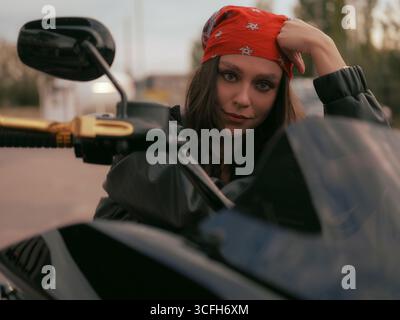 Jeune femme motard élégante en veste en cuir et bandana rouge est assis en toute confiance sur une moto, regardant la caméra. Urbain cool, dynamique et rebelle Banque D'Images