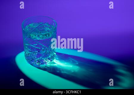 Une photo d'un verre de whisky coupé rempli d'eau, éclairé par un rétro-éclairage bleu, projetant des motifs caustiques sur le sol en raison de sa forme et de sa lumière refr Banque D'Images