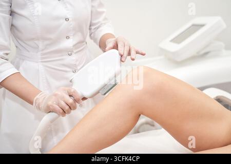 Technicien féminin effectuant l'épilation au laser sur la jambe de femme adulte caucasienne en milieu clinique. Banque D'Images