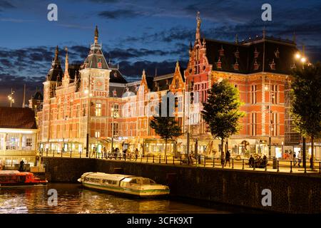 Amsterdam Centraal Station illuminée au crépuscule avec un bateau sur le canal, pays-Bas Banque D'Images