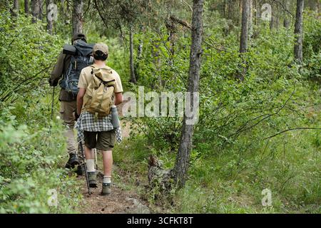 Homme d'âge moyen et garçon marchant ensemble à travers le sentier forestier transportant des sacs à dos et des cannes à pêche, marchant côte à côte entouré d'arbres verts denses Banque D'Images