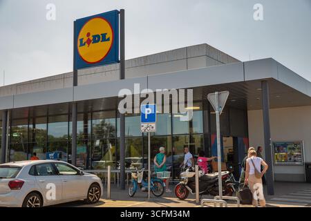 Porec, Croatie - 31 mai 2025 : façade moderne de l'épicerie Lidl avec logo bleu et jaune distinctif. Acheteurs entrant dans le magasin avec des voitures garées, bicyc Banque D'Images