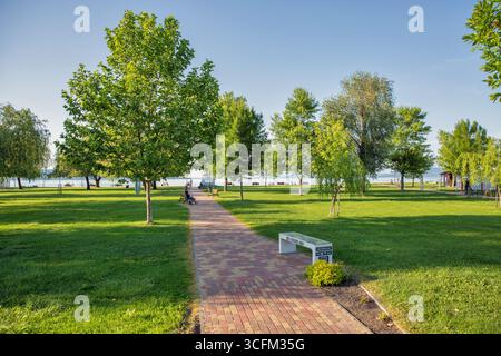 Balatonmariafurdo, Hongrie - 31 mai 2025 : pittoresque parc central public avec une passerelle pavée de briques menant à travers une pelouse verdoyante vers le lac, les arbres Banque D'Images