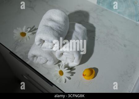 Les serviettes blanches roulées sont soigneusement empilées sur un comptoir en marbre, accompagnées de marguerites fraîches. Un canard en caoutchouc jaune joyeux ajoute une touche ludique à cette atmosphère de spa tranquille. Banque D'Images