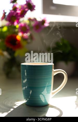 Une tasse de café fumant se trouve sur un comptoir, entouré de fleurs colorées en arrière-plan. La lumière vive du soleil filtre à travers la fenêtre, créant une atmosphère chaleureuse et accueillante. Banque D'Images