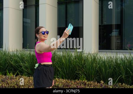 Une femme latine souriante dans un haut de sport rose et un short noir prend un selfie avec son smartphone à l'extérieur. Bâtiment moderne et verdure dans le backgrou Banque D'Images
