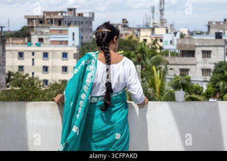 Femme bengali dans un saree turquoise se tient sur un toit, ses longs cheveux tressés en cascade dans son dos. Elle regarde la ville avec une tasse à café à côté Banque D'Images