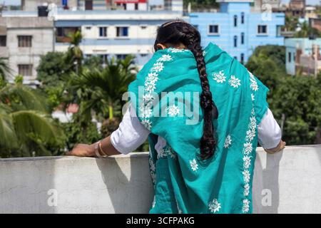 Femme bengali dans un saree turquoise, ses longs cheveux tressés visibles de l'arrière, se tient sur un toit regardant vers le bas. Banque D'Images