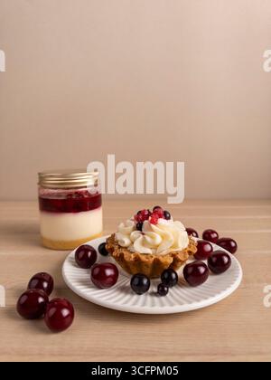 Une petite tarte avec de la crème fouettée et des baies sur une assiette blanche, entourée de cerises. En arrière-plan, un pot de dessert en couches avec des fruits, de la crème, un Banque D'Images