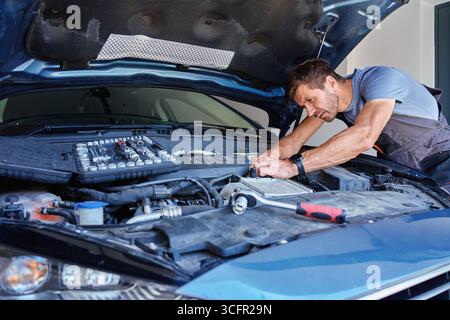 Mécanicien masculin utilisant une clé à douille pour réparer le moteur de voiture dans le garage. Homme avec des outils de réparation dans les mains sous le capot ouvert Banque D'Images