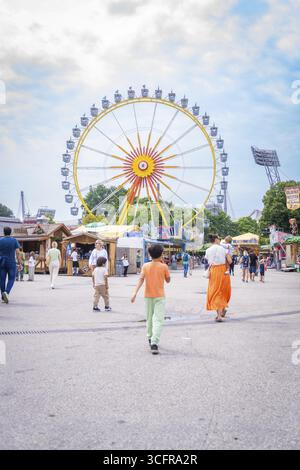 Les gens sur une place dans un parc d'attractions avec une grande roue en arrière-plan, Olympiapark, Munich, Allemagne Banque D'Images