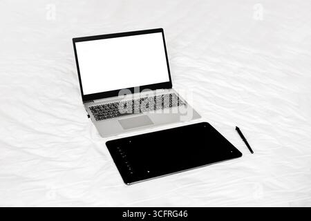 ordinateur portable ouvert avec écran vierge et tablette graphique noire avec stylo sur un lit blanc. tablette pour dessin, maquette. copier l'espace Banque D'Images