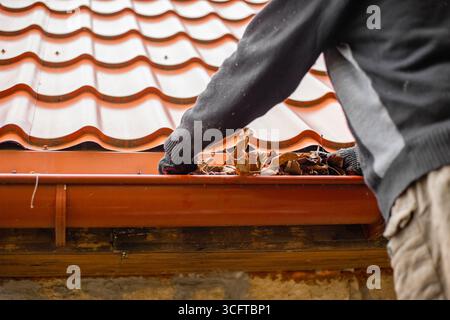 Un homme portant des gants enlève les feuilles mortes et les débris d'une gouttière sous un toit de tuiles métalliques rouges. Banque D'Images
