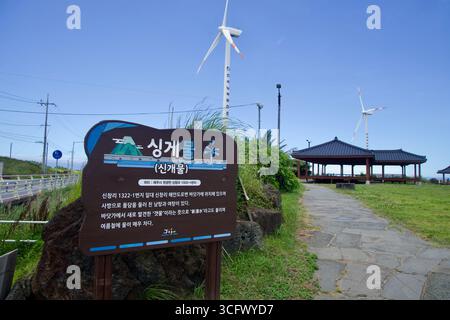 Le panneau d’information Singgemul se trouve à côté d’un pavillon traditionnel alors que les éoliennes tournent au-dessus de Sinchang Windmill Coastal Road sur la côte ouest de Jeju. Banque D'Images