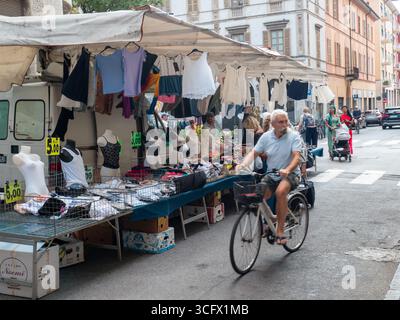 Cremona, Italie- 23 août 2025 homme âgé fait des cycles devant l'étal de marché en plein air dans la ville italienne historique avec des vêtements et des accessoires exposés pour sal Banque D'Images