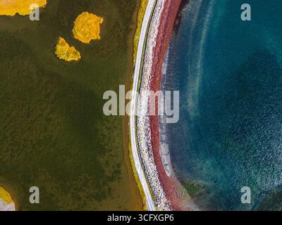 Vue aérienne d'une route traversant des paysages contrastés, où les eaux sombres rencontrent le rivage rouge, Kálfshamarsvík, Húnabyggð, Islande. Banque D'Images