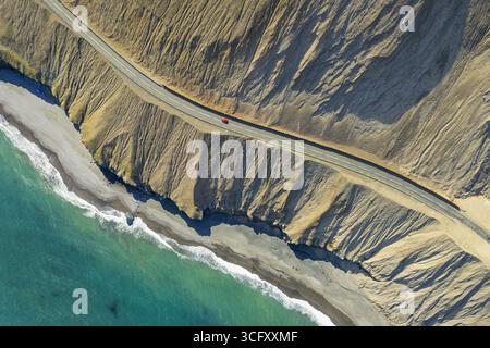 Vue aérienne d'une voiture rouge solitaire navigue sur une route côtière encadrée par des falaises accidentées et ensoleillées et l'étreinte turquoise de la mer, les routes islandaises, l'Islande. Banque D'Images