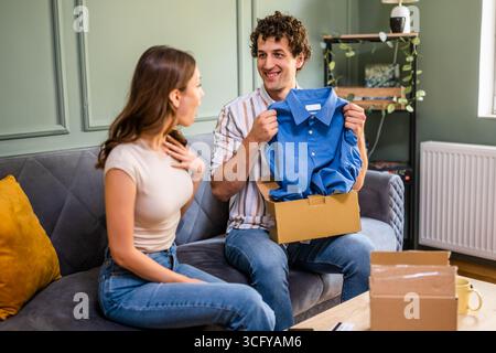 Couple adulte déballage leurs colis livrés à domicile. L'homme montre sa nouvelle chemise à sa femme. Banque D'Images