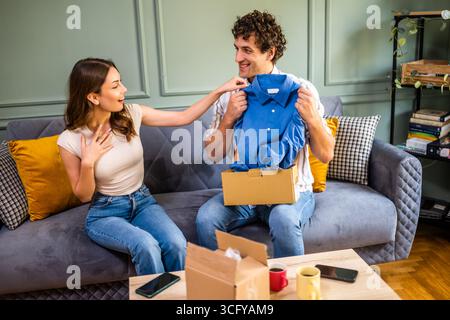 Couple adulte déballage leurs colis livrés à domicile. L'homme montre sa nouvelle chemise à sa femme. Banque D'Images