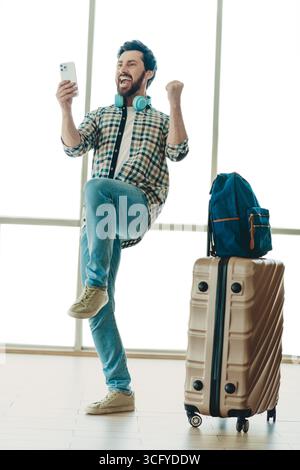 Voyageur excité célébrant la victoire au terminal de l'aéroport, tenant smartphone, à côté de la valise et sac à dos, émotions vibrantes Banque D'Images